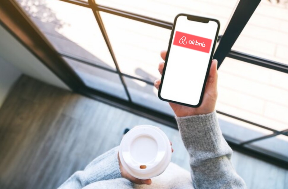 a woman in a sweater holding a cup of coffee and a smartphone with the Airbnb logo on the screen