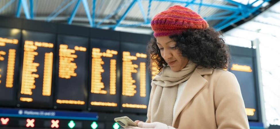 a woman wearing a rat beret, beige coat, scarf, and gloves holding a smartphone in the airport